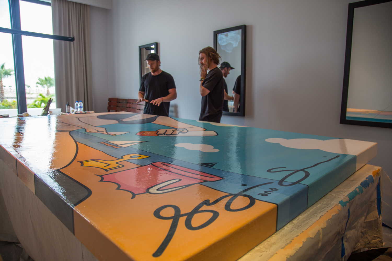 Two men standing indoors beside a large colorful painting on a table, one man gesturing while the other listens thoughtfully.