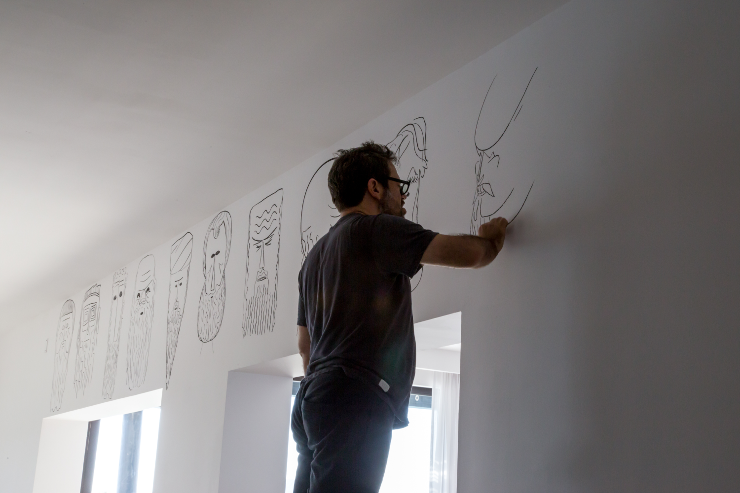 Man drawing abstract faces with long beards on a white wall near a window.