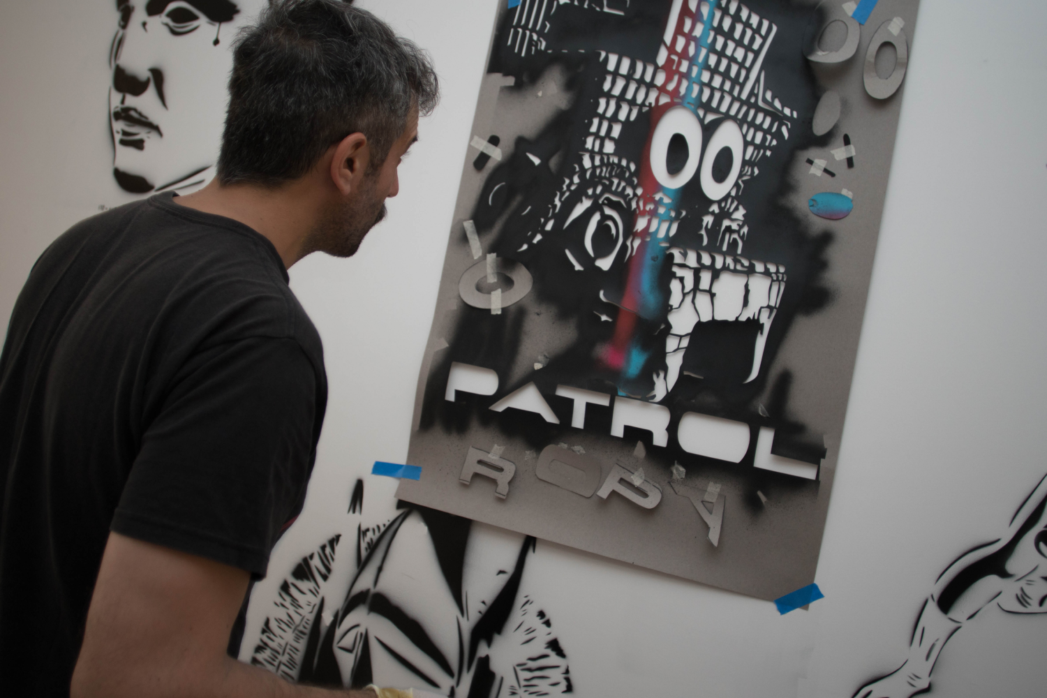 Man with dark hair and beard looks at graffiti art stencil of a cartoonish skull with wide eyes and the text 'PATROL' on a white wall.