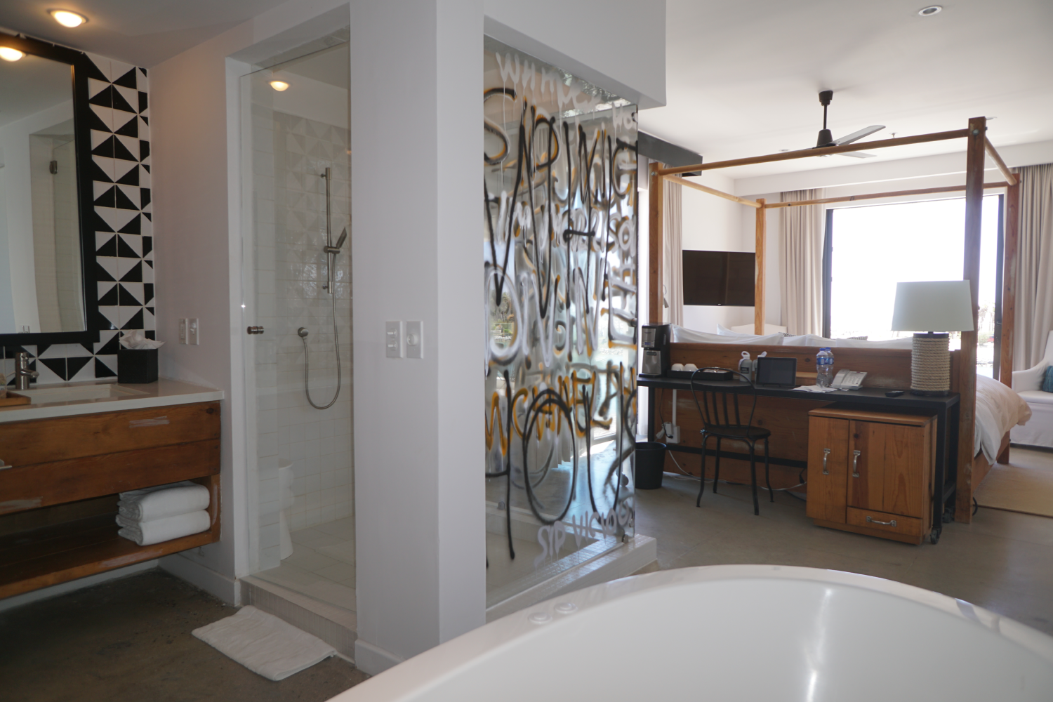Modern hotel room with a glass-walled shower, wooden vanity, round bathtub, wooden four-poster bed, and desk with coffee maker and lamp.