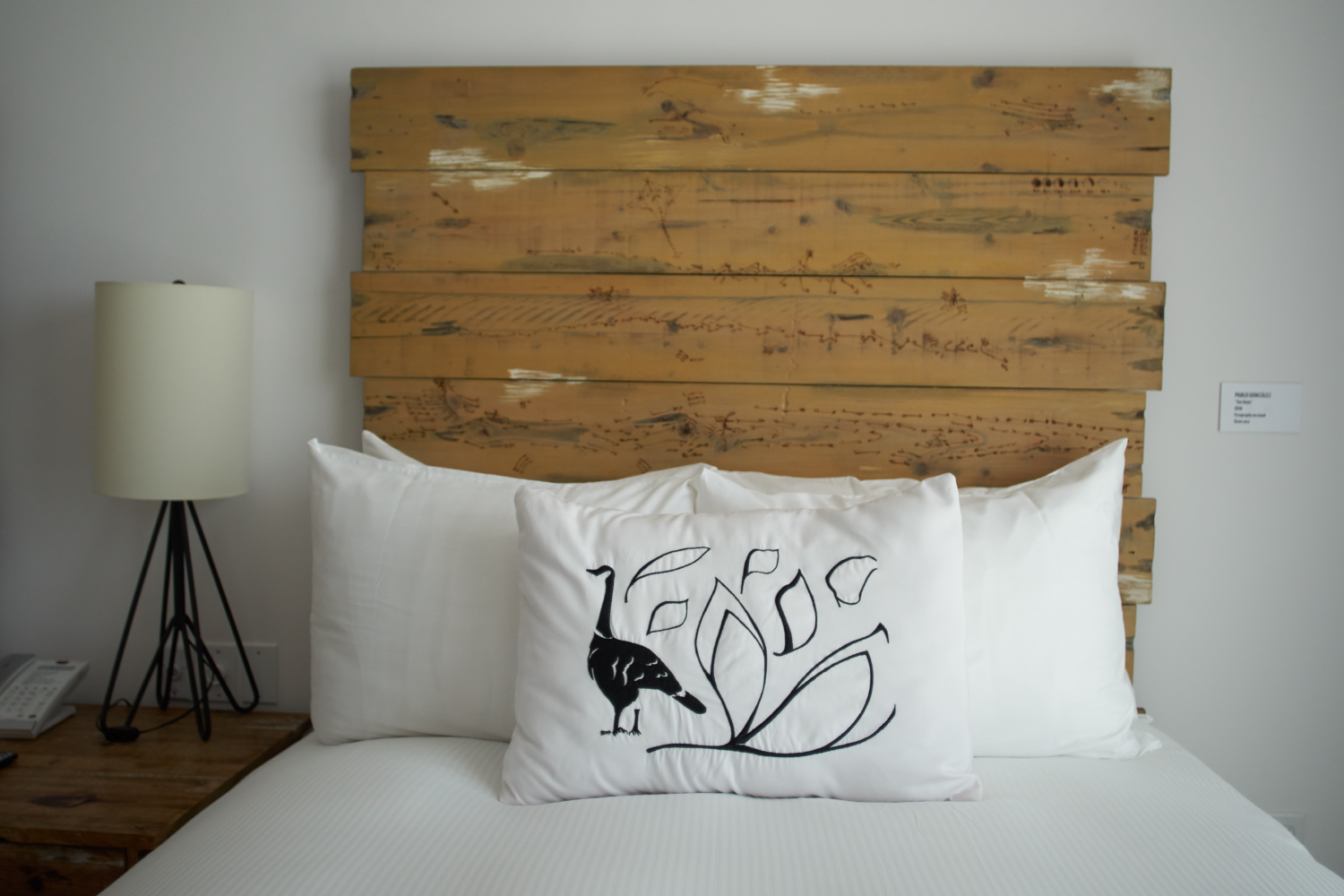 White bed with a rustic wooden headboard, featuring white pillows and a decorative pillow embroidered with a black bird and abstract leaf design.