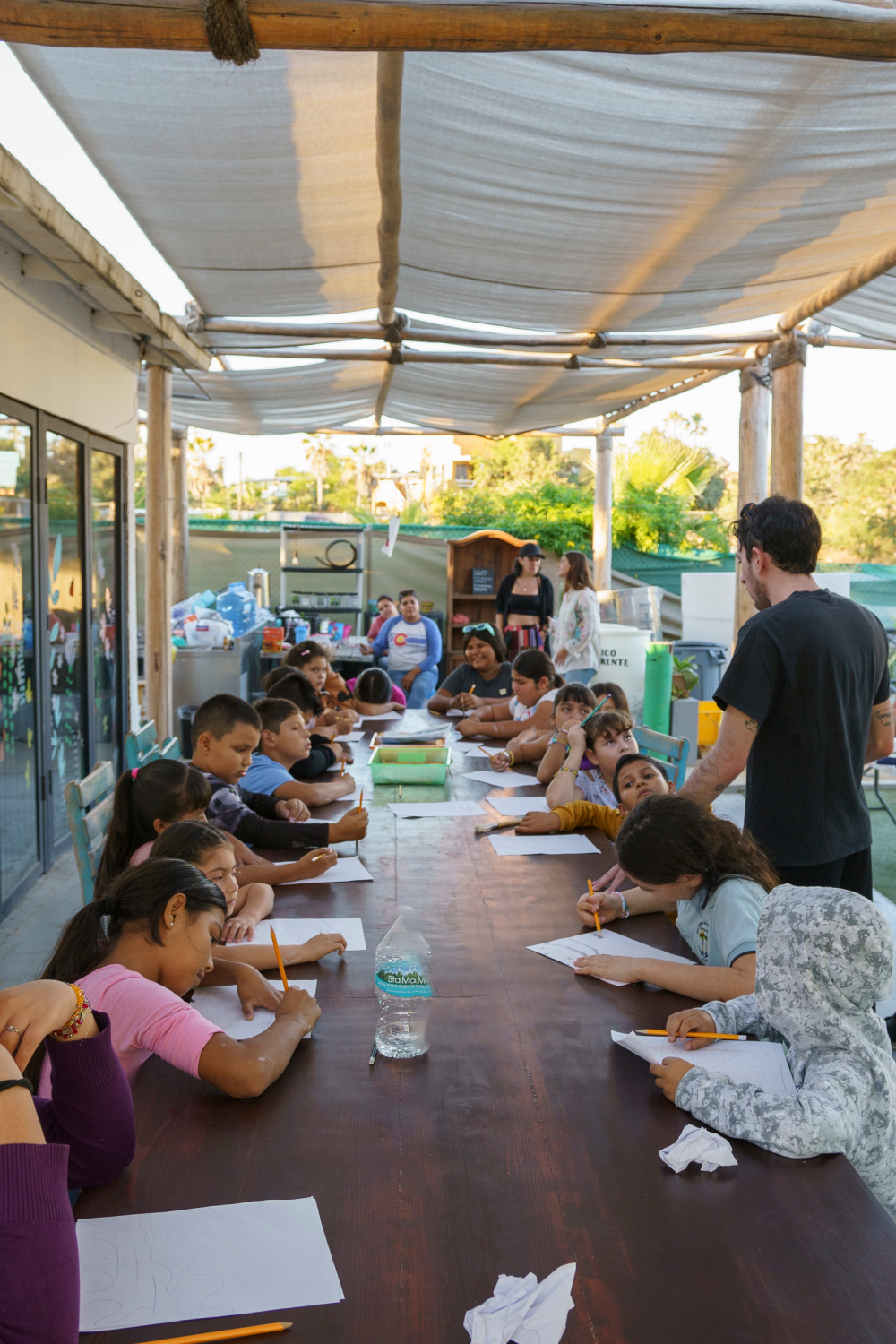 Group of children seated around a long table outdoors under canopy, drawing and writing on paper with pencils while a man stands nearby.