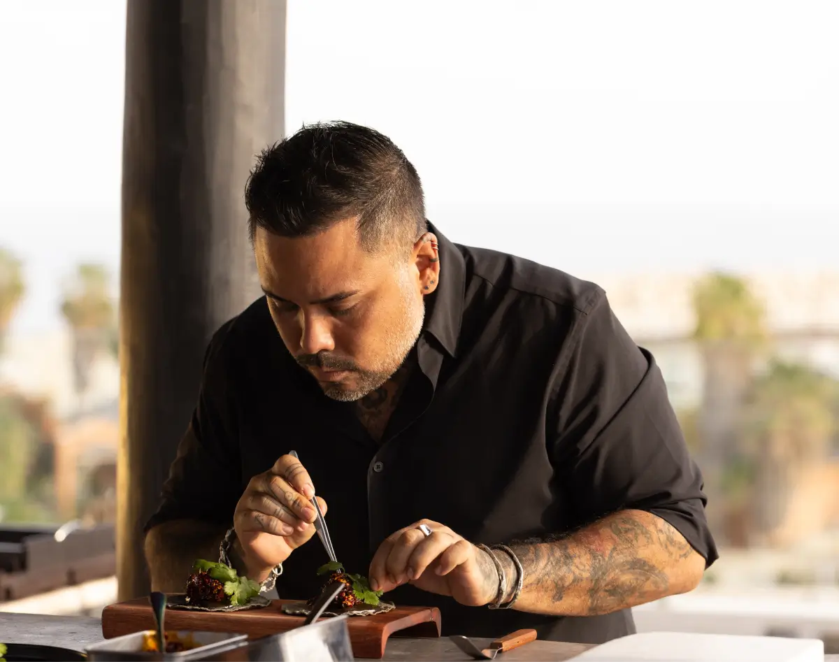 Chef con tatuajes y camisa negra decorando cuidadosamente la comida en una tabla de madera con unas pinzas.