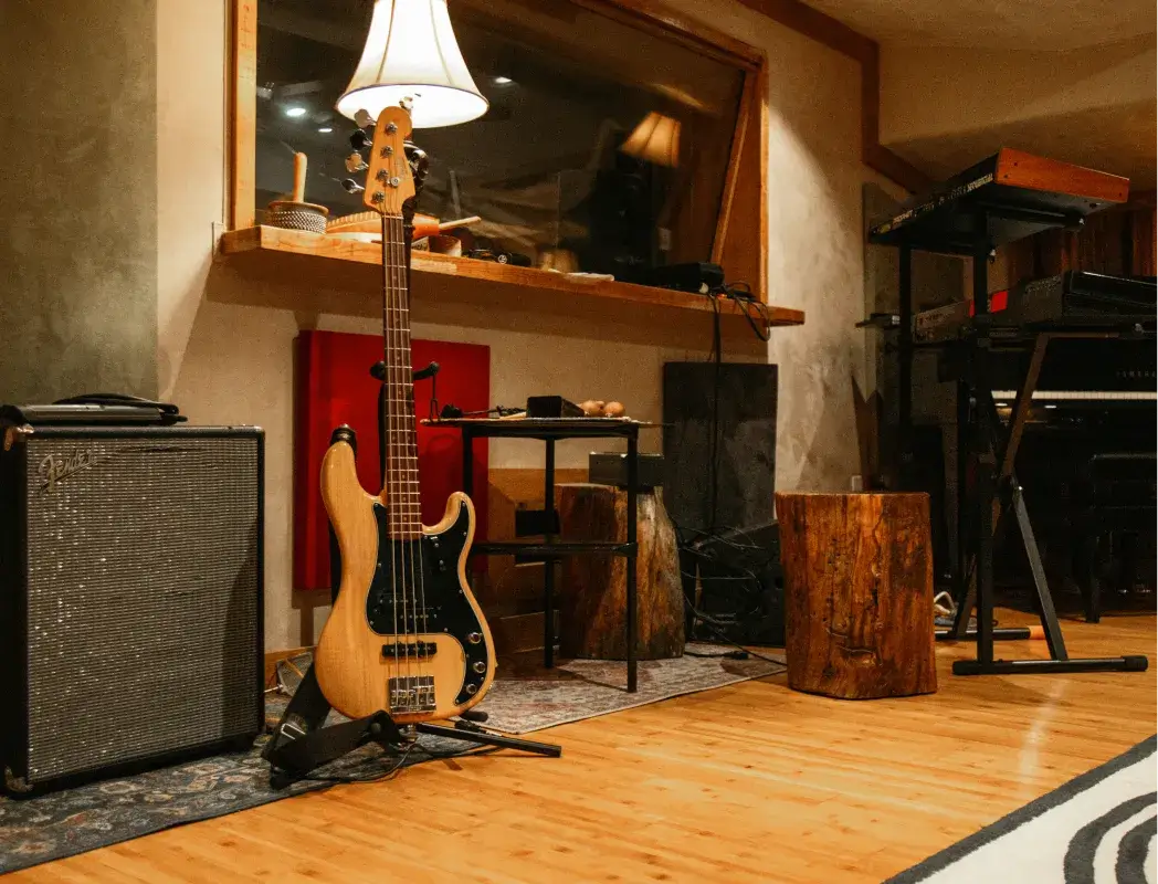 Light wood bass guitar on a stand next to a Fender amplifier in a music studio with wooden floor and recording equipment.