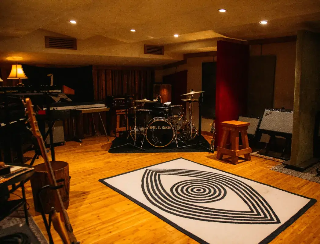 Cozy music studio with drum set labeled 'Hotel El Ganzo,' keyboards, guitar, wooden stool, and a distinctive black and white eye-patterned rug on wooden floor.