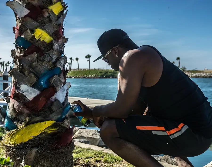 Hombre con camiseta sin mangas negra y gorra pintando con spray manchas de colores en una palmera cerca de la costa.