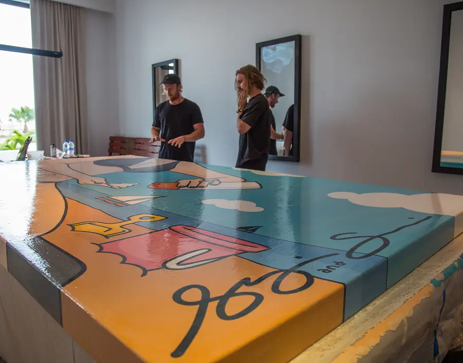 Dos hombres de pie en el interior junto a un gran cuadro colorido sobre una mesa, uno gesticulando mientras el otro escucha pensativo.