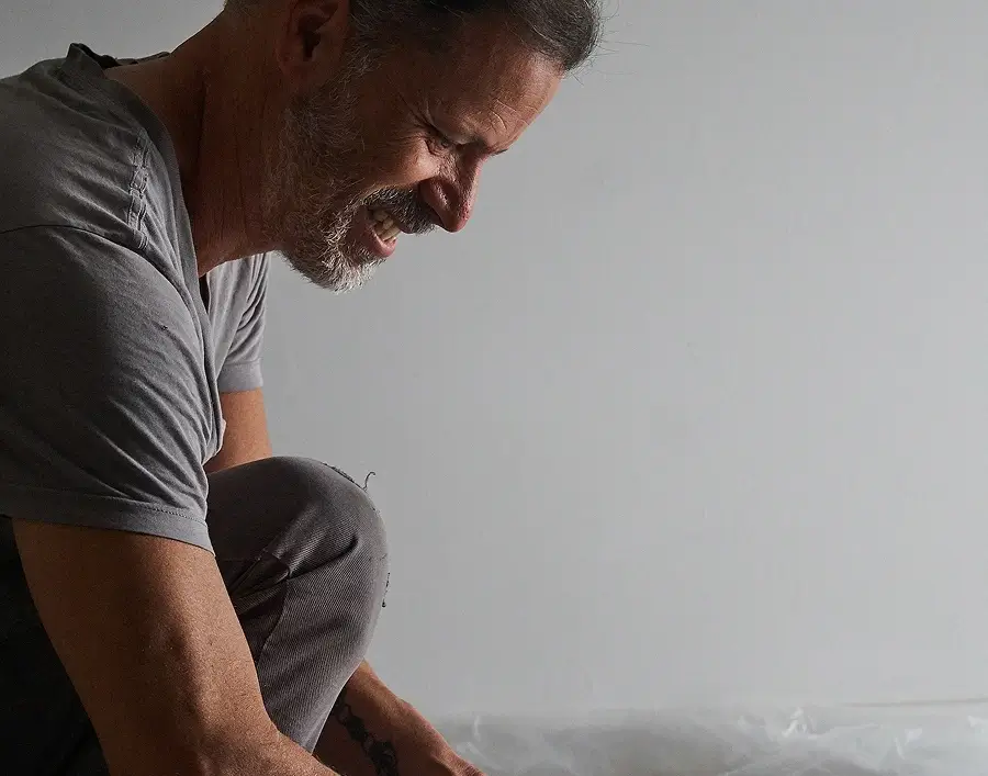 Hombre sonriente arrodillado y abriendo un cubo blanco con una etiqueta azul sobre un suelo cubierto de plástico.
