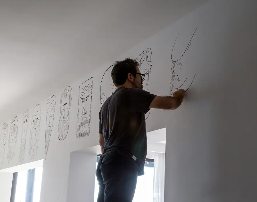 Hombre dibujando caras abstractas con largas barbas en una pared blanca cerca de una ventana.