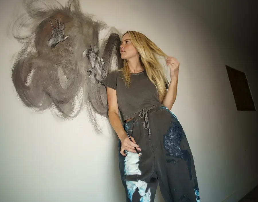 Mujer rubia sosteniendo un pincel y levantándose el pelo, de pie frente a una pared con un oscuro dibujo abstracto pintado a mano.