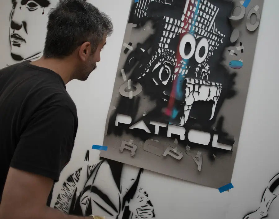 Un hombre con cabello oscuro y barba mira una plantilla de arte grafiti de una calavera caricaturesca con ojos grandes y el texto «PATROL» en una pared blanca.