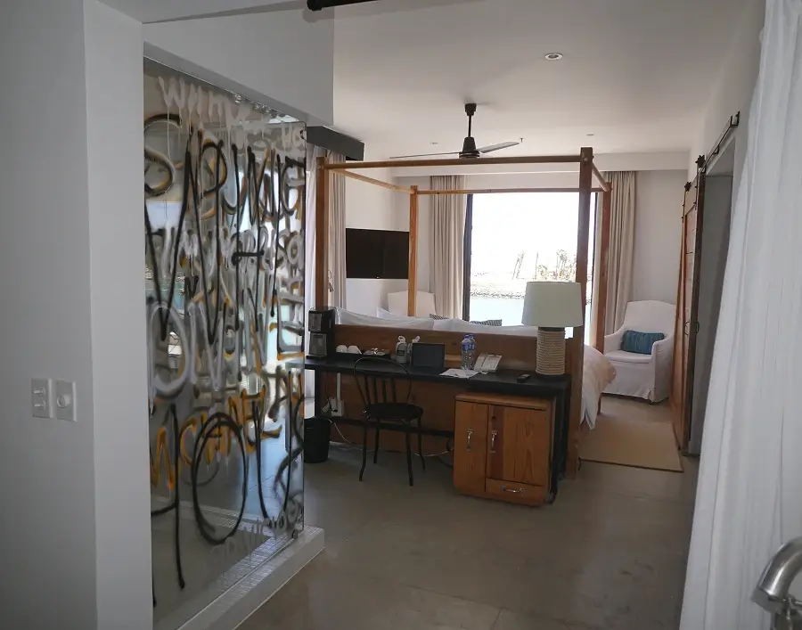 Habitación de hotel moderna con ducha con paredes de cristal, tocador de madera, bañera redonda, cama con dosel de madera y escritorio con cafetera y lámpara.