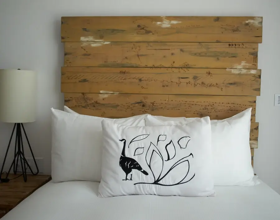 Cama blanca con cabecero rústico de madera, almohadas blancas y una almohada decorativa bordada con un pájaro negro y un diseño abstracto de hojas.