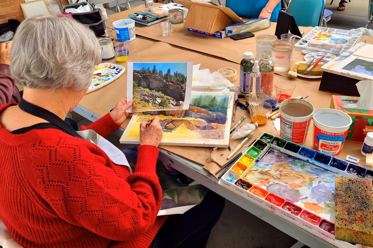 Spokane watercolor society member in a red sweater painting a landscape with rocks and trees using watercolors, referencing a photograph.