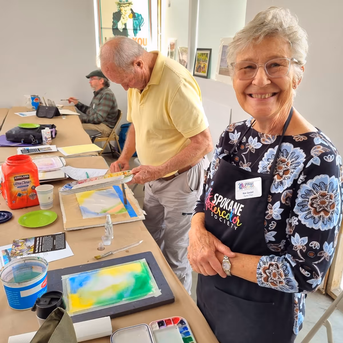 spokane watercolor society president standing wearing glasses and a Spokane Watercolor Society apron stands next to a man painting at a table with watercolor supplies.