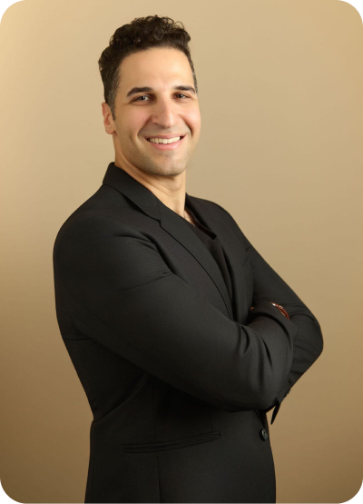 Smiling man with dark curly hair in a black blazer standing with arms crossed against a beige background.