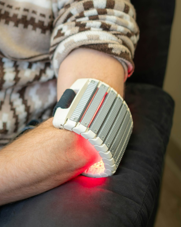 Person wearing a red and white light therapy device on their elbow while resting on a black surface.