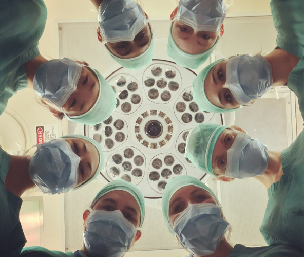Eight surgeons wearing surgical masks and caps looking down at the camera under an operating room light.