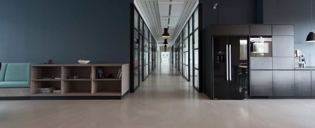 Modern office hallway with glass-walled rooms on both sides, pendant lights hanging from the ceiling, a built-in shelf with books and a bowl, and a kitchen area with black cabinets and appliances.
