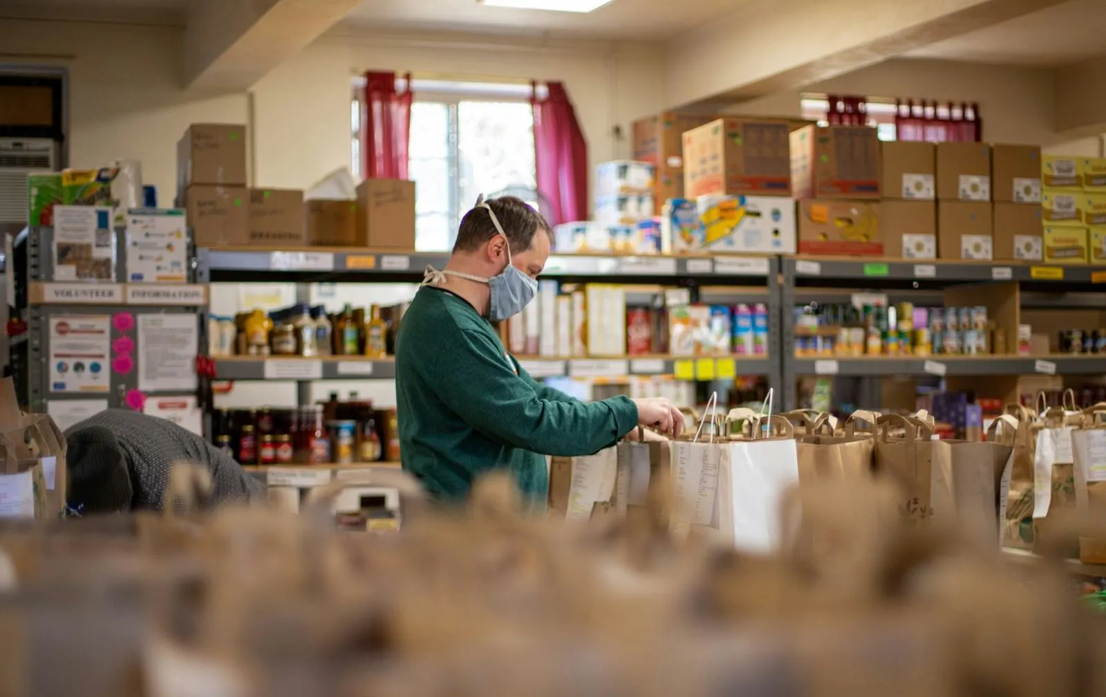 A man wearing a mask and green sweater packs paper bags in a food pantry, surrounded by shelves of canned goods and boxes. The mood is focused and industrious.