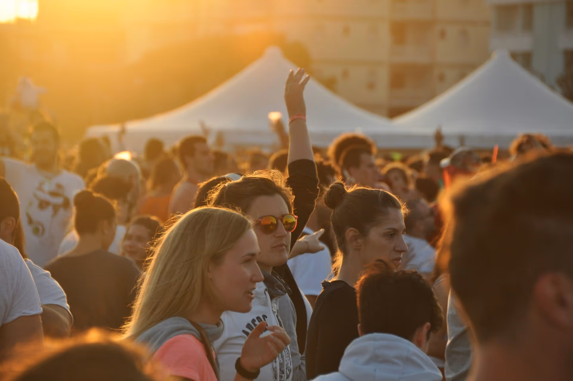 A vibrant outdoor festival at sunset with people gathered in front of white tents. Warm sunlight creates a lively and energetic atmosphere.