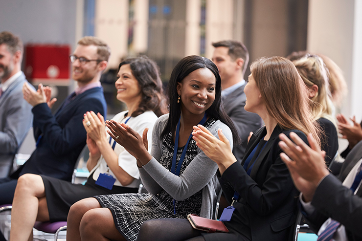 A diverse group of people sitting in rows clapping and smiling during a professional event or conference.