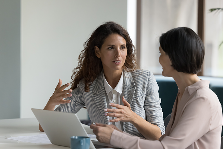 Two professional women discussing something at a desk with an open laptop and documents.