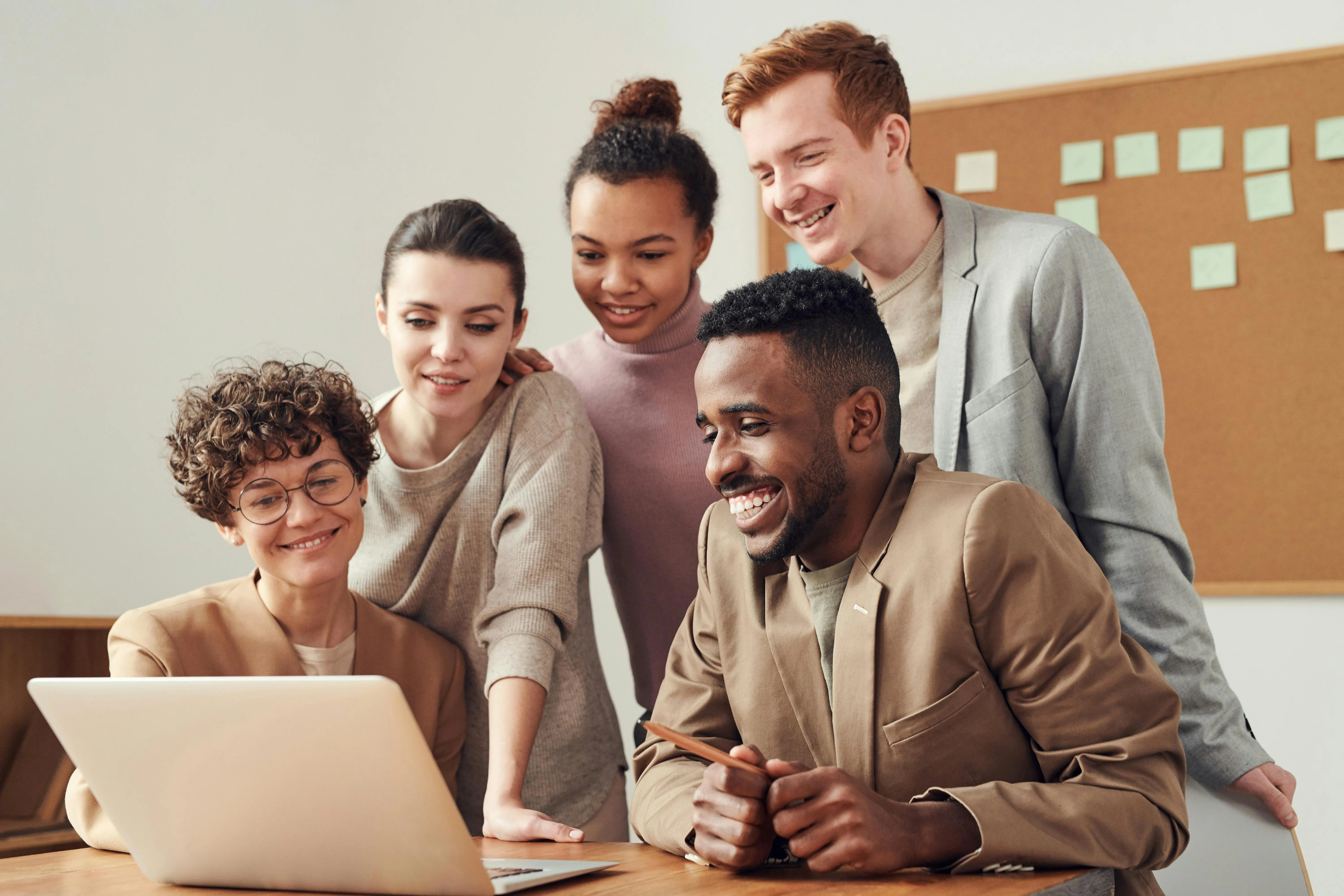 A happy, diverse team of professionals gathered around a laptop, smiling and collaborating on IT solutions in a modern office setting.