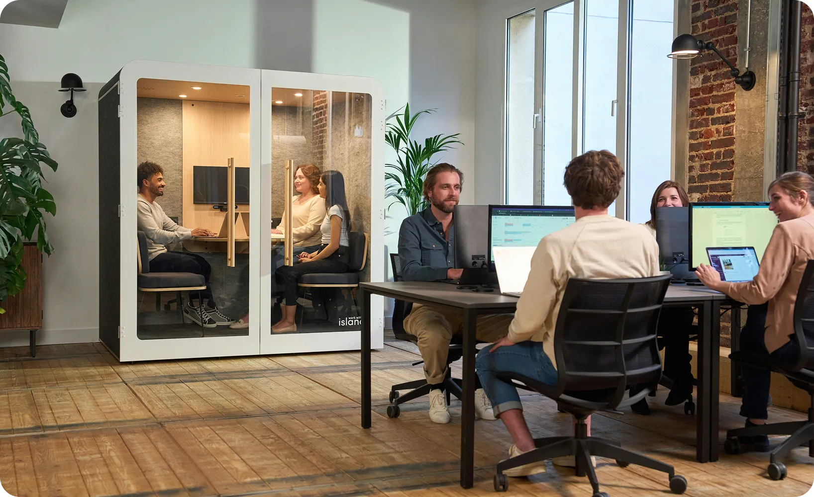 A wide view of a modern, open-plan office featuring a diverse team of professionals, with four people working at a shared collaborative desk in the foreground and three others having a meeting inside a soundproof, glass-walled acoustic pod.