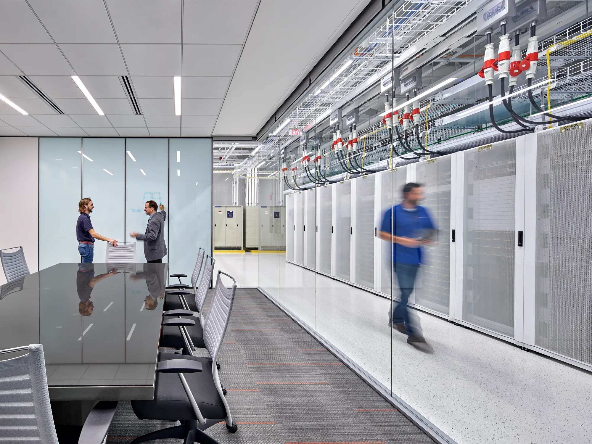 A modern corporate conference room adjacent to a high-density data center, separated by glass walls, featuring white server racks and overhead power cabling to illustrate advanced infrastructure design.