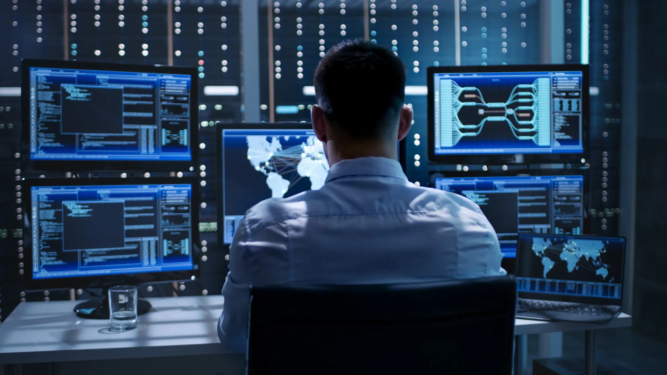 A network engineer viewed from behind, monitoring multiple screens displaying global connectivity maps, system code, and data diagrams in a high-security network operations center (NOC).
