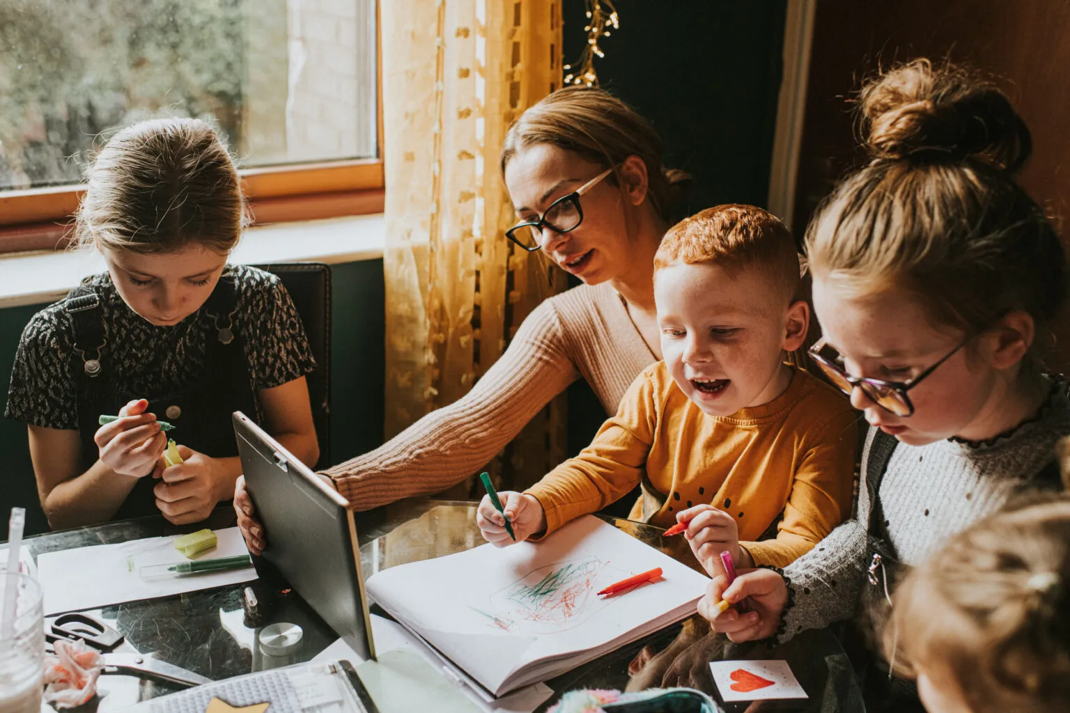 A teacher or parent sitting at a table with three young children, helping them engage in creative drawing and remote learning using a tablet in a well-lit home or classroom setting.