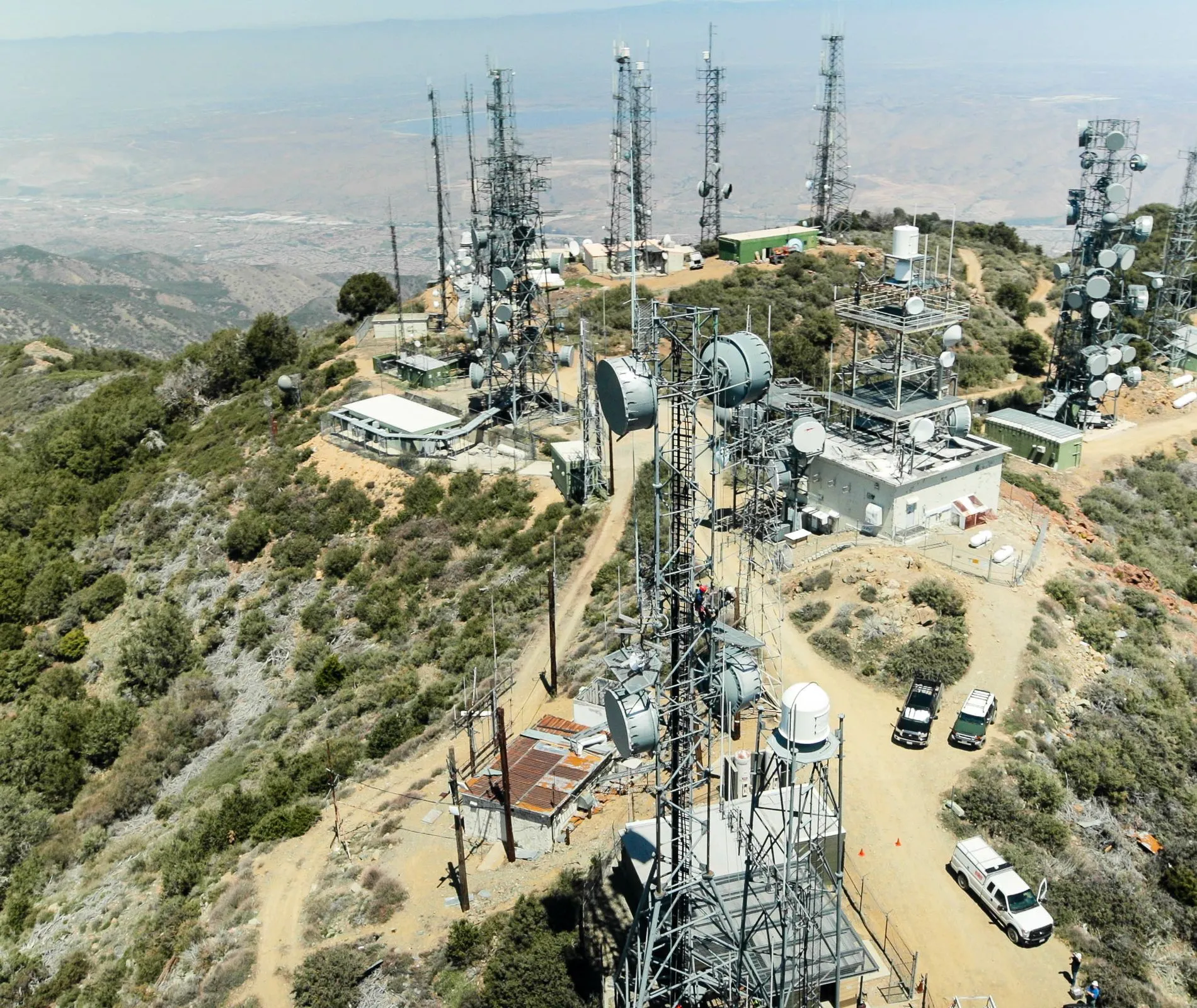 An aerial view of a remote mountaintop telecommunications site featuring multiple lattice towers equipped with microwave dishes and antennas, providing critical wireless coverage and infrastructure for the surrounding region.