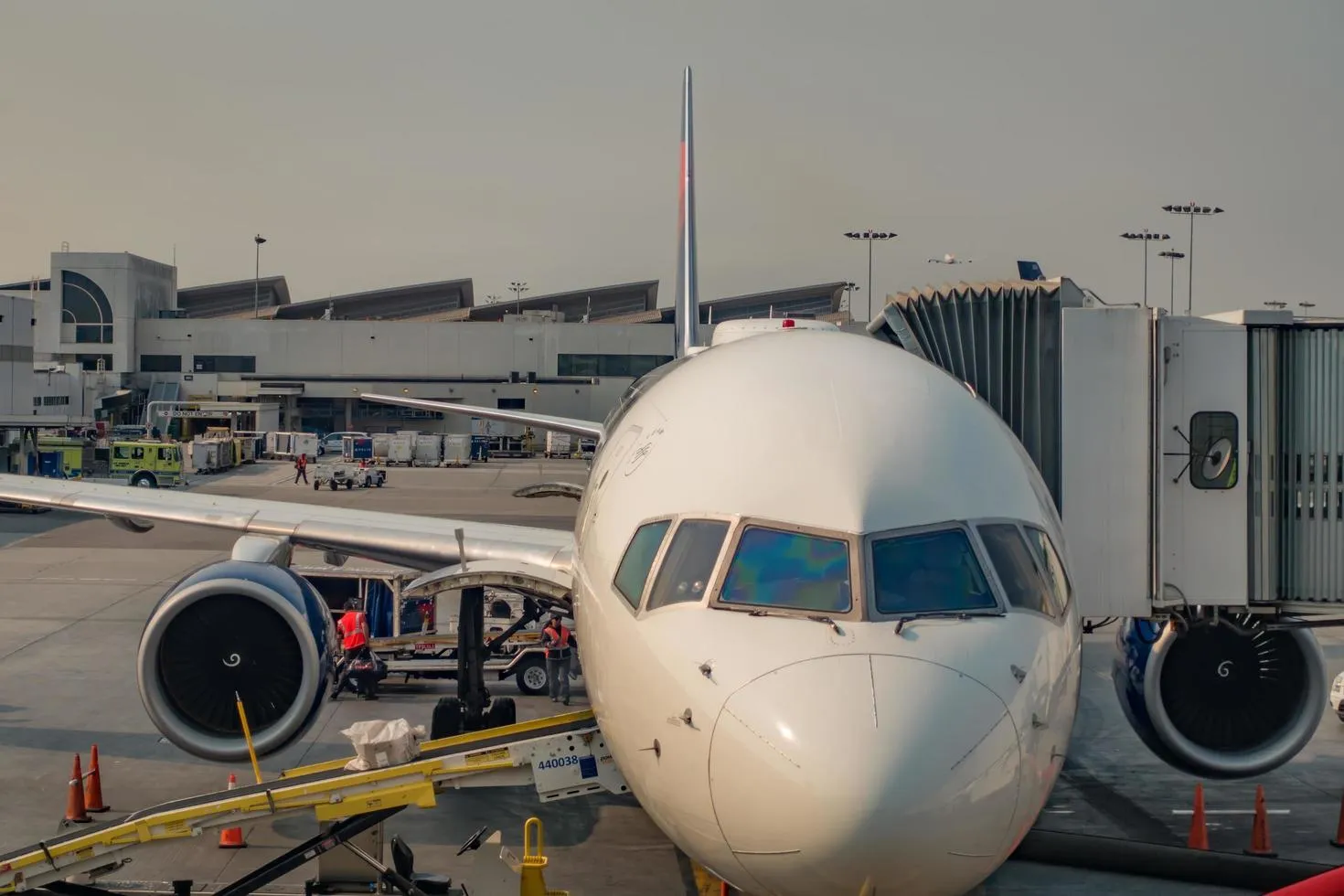 A close-up, front-facing view of a commercial airliner at an airport gate, with ground crew loading cargo and a jet bridge connected, representing aviation infrastructure and ground support services.