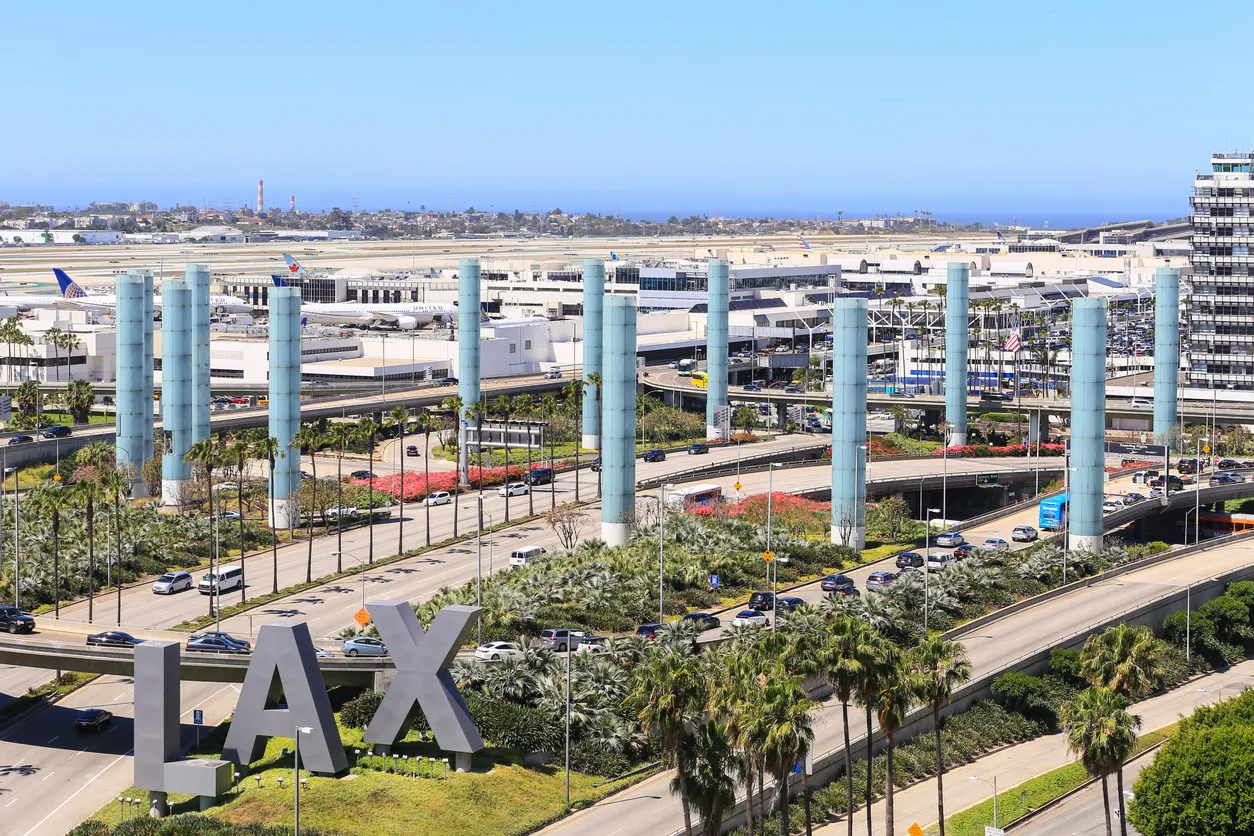 A wide aerial view of the Los Angeles International Airport (LAX) entrance, featuring the iconic light pylons, the large 'LAX' sign, and terminal traffic infrastructure under a clear blue sky.