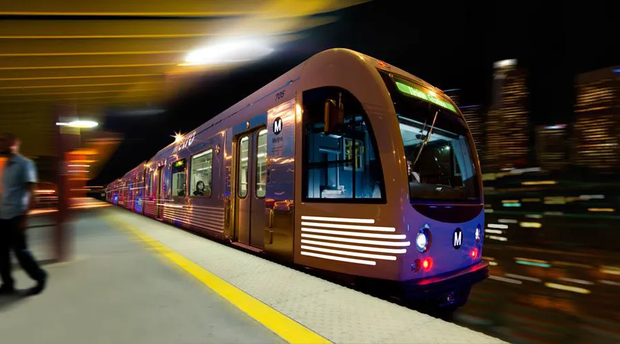 A motion-blurred view of an LA Metro train at a station platform at night, with city lights and long-exposure streaks representing urban transit efficiency.