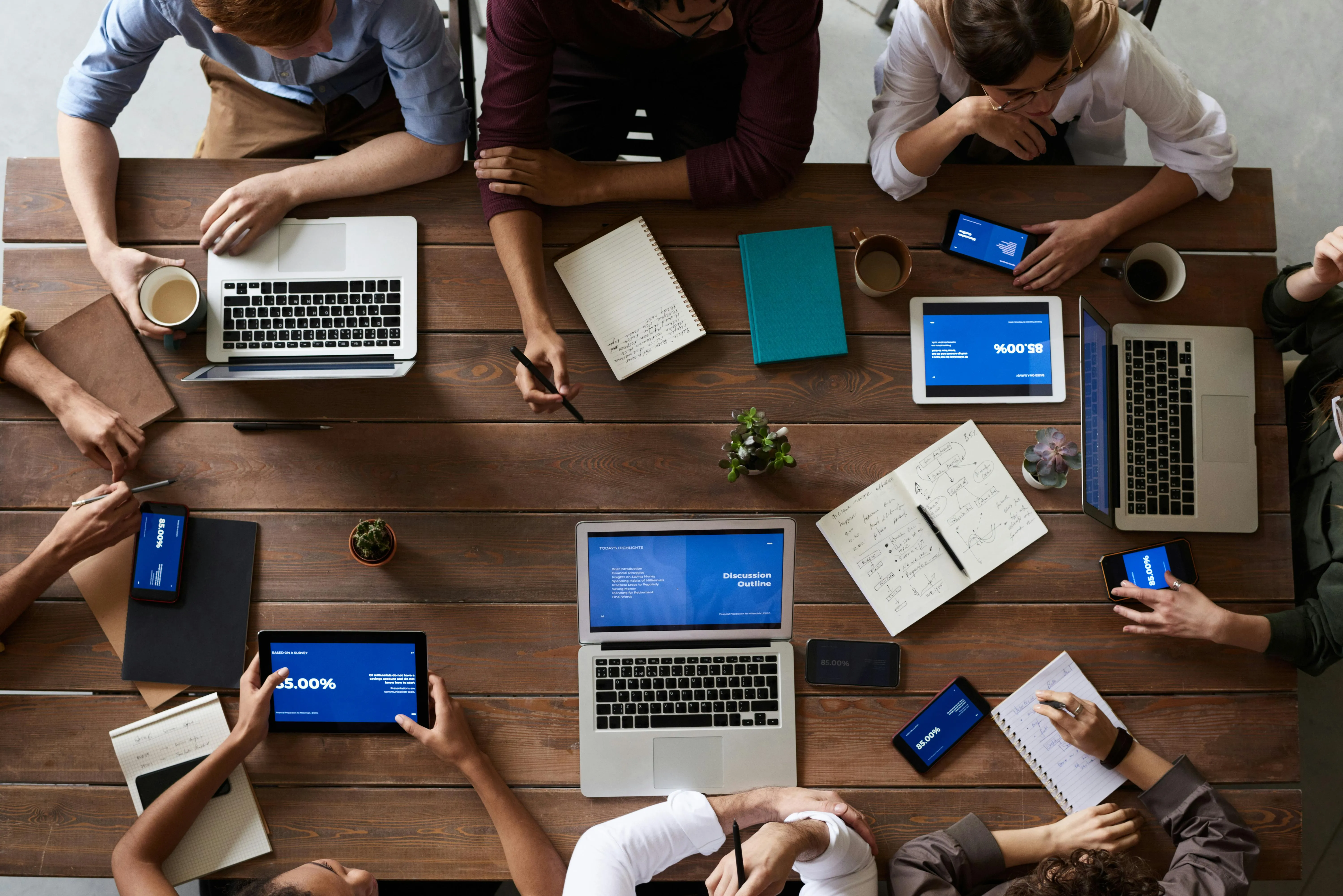 A close-up of a diverse group of four professionals—two women and two men—collaborating around a small, circular table. They are reviewing printed documents, writing, and looking at a tablet together in a modern, light-filled office.