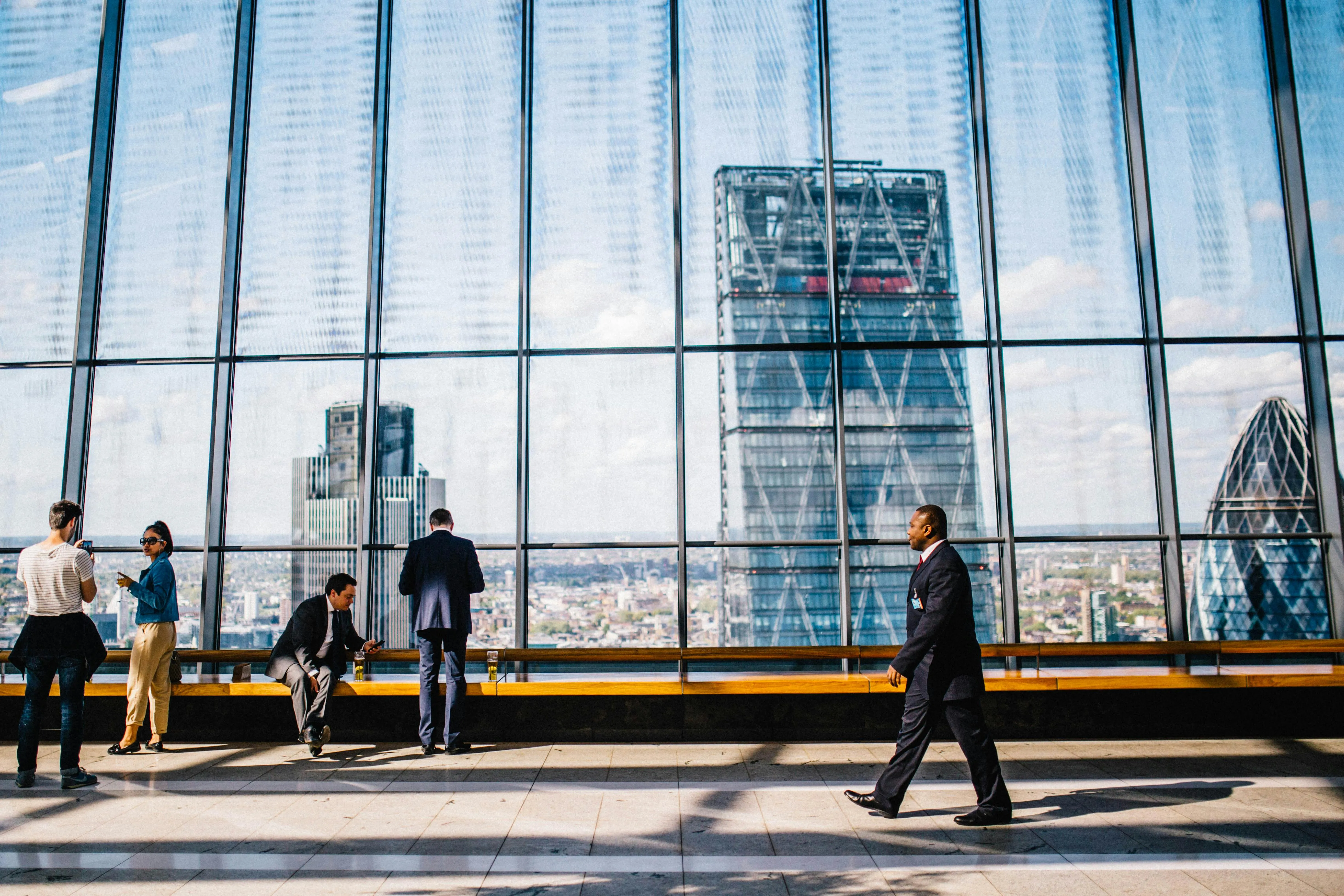 Professionals and visitors standing inside a modern corporate high-rise with floor-to-ceiling glass windows, looking out over a panoramic view of the city skyline, featuring iconic skyscrapers.