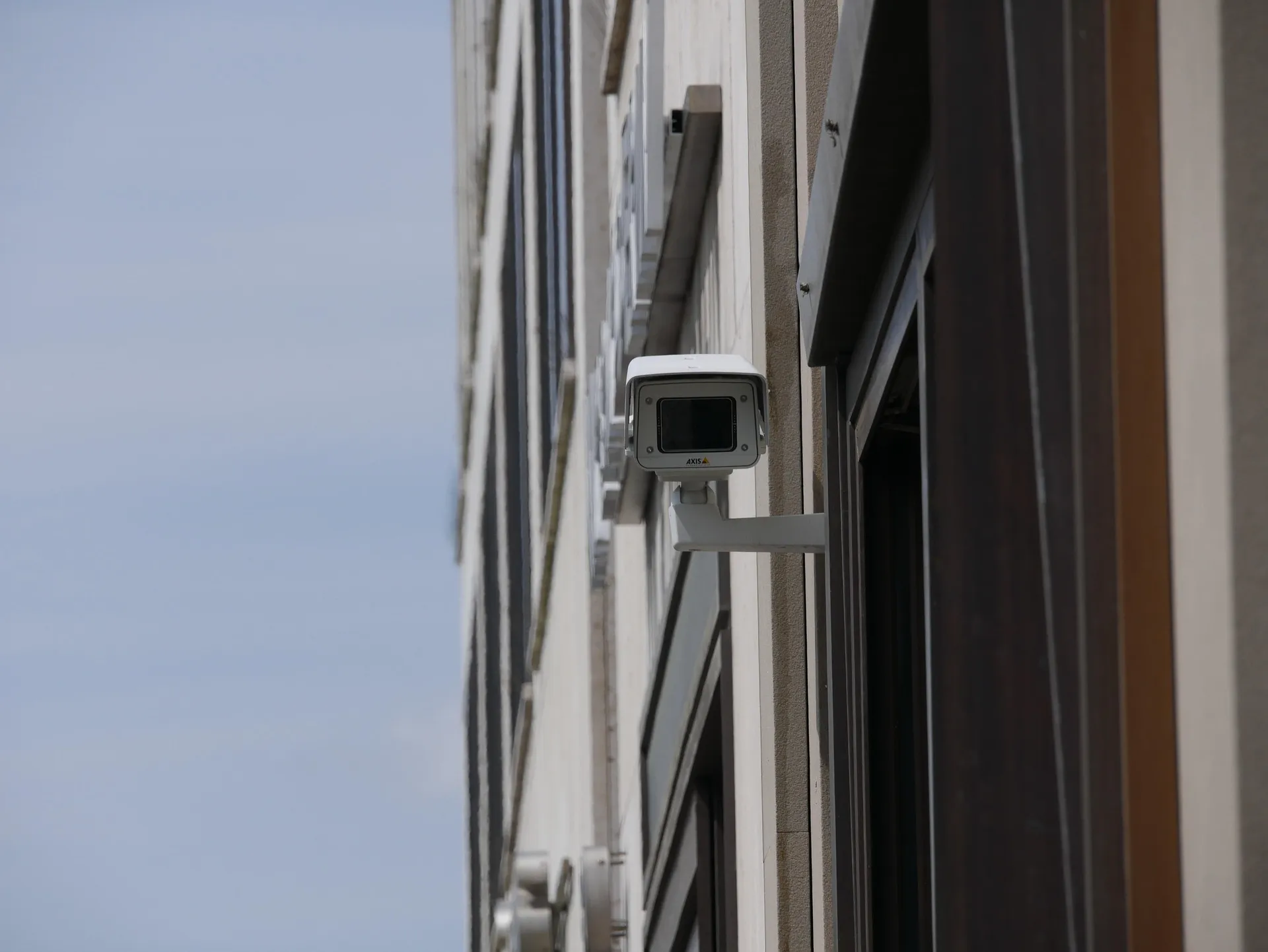 A white Axis-branded security camera mounted to the side of a tall office building, providing surveillance of the surrounding urban area.
