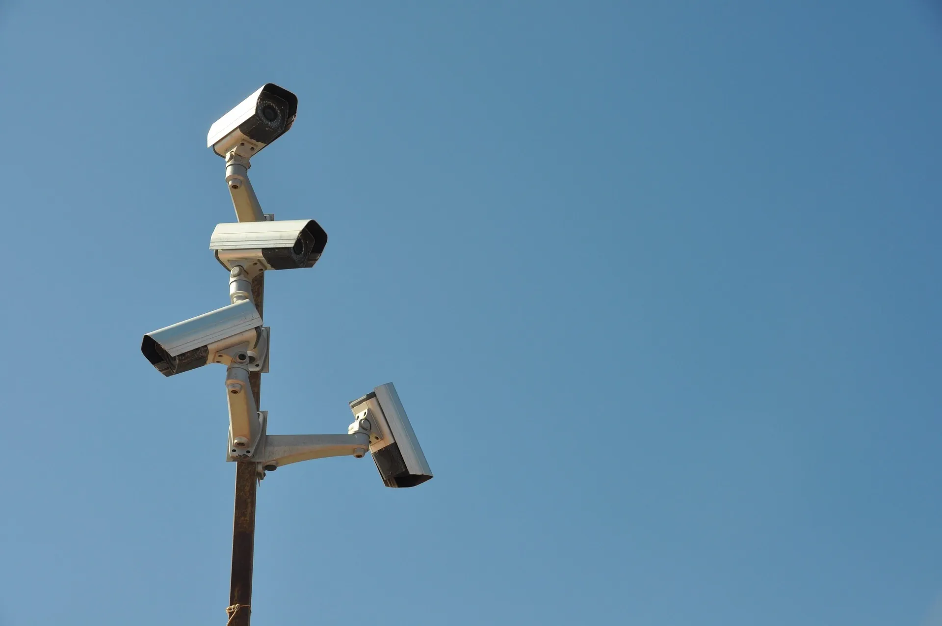 Four white professional CCTV security cameras mounted in different directions on a single pole against a clear blue sky, illustrating 360-degree site surveillance and monitoring.
