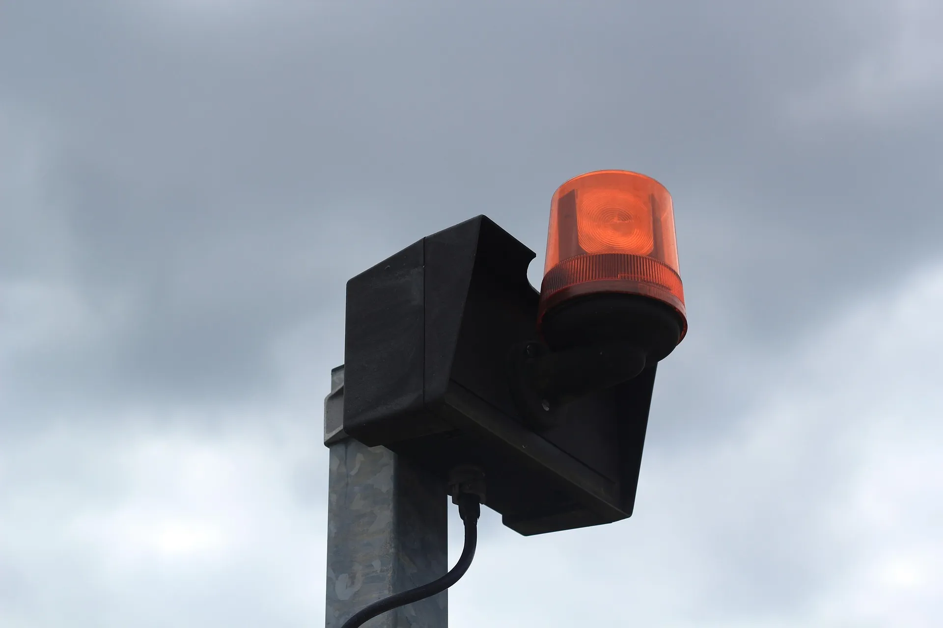 A close-up of an industrial orange warning beacon and alarm siren mounted on a pole against a cloudy sky, used for safety signaling at a project site.