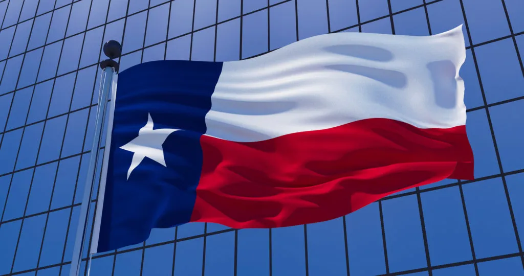 The state flag of Texas waving prominently in front of a modern blue glass office building, symbolizing E.K. Associates' operations and project management in the Texas region.