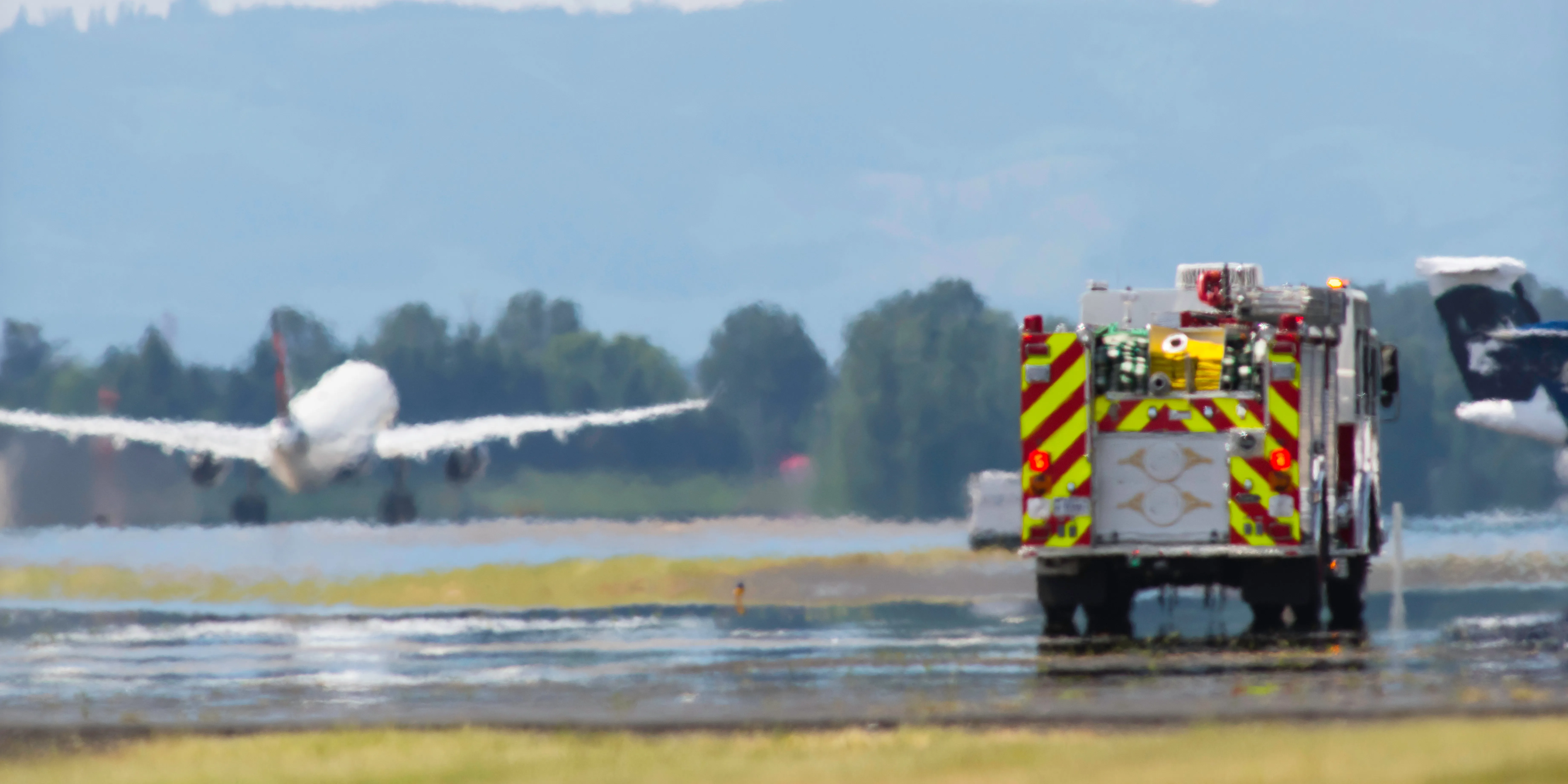 An airport fire and rescue truck positioned on a runway as a commercial aircraft takes off in the background, representing aviation safety protocols and emergency response infrastructure.