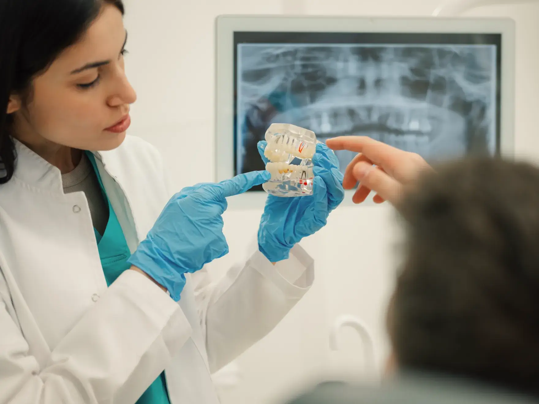 Dentist wearing blue gloves explaining a dental model to a patient, with a dental X-ray displayed in the background.