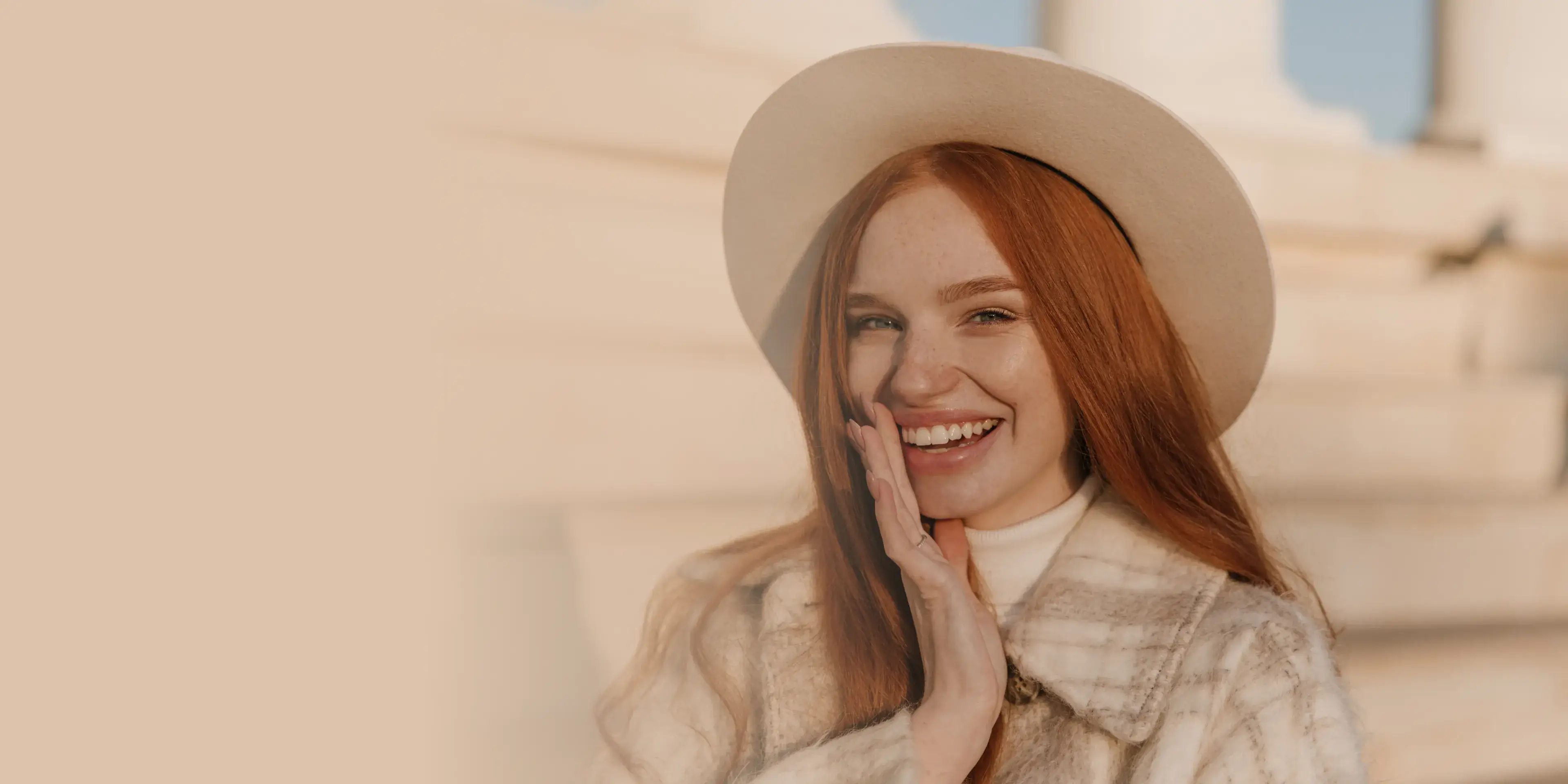 Smiling young woman with red hair wearing a beige hat and plaid coat, posing outdoors.