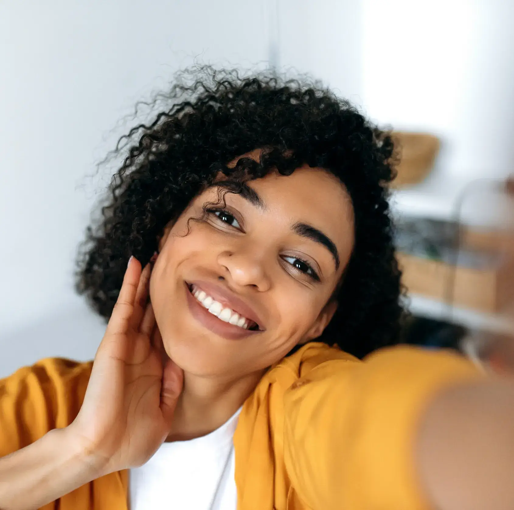 Smiling young woman with curly hair wearing a yellow jacket taking a selfie indoors.
