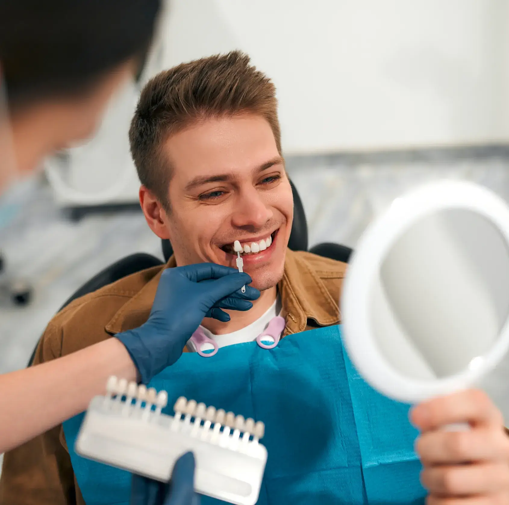 Dentist holding a teeth shade guide next to a smiling man's teeth as he looks into a handheld mirror.