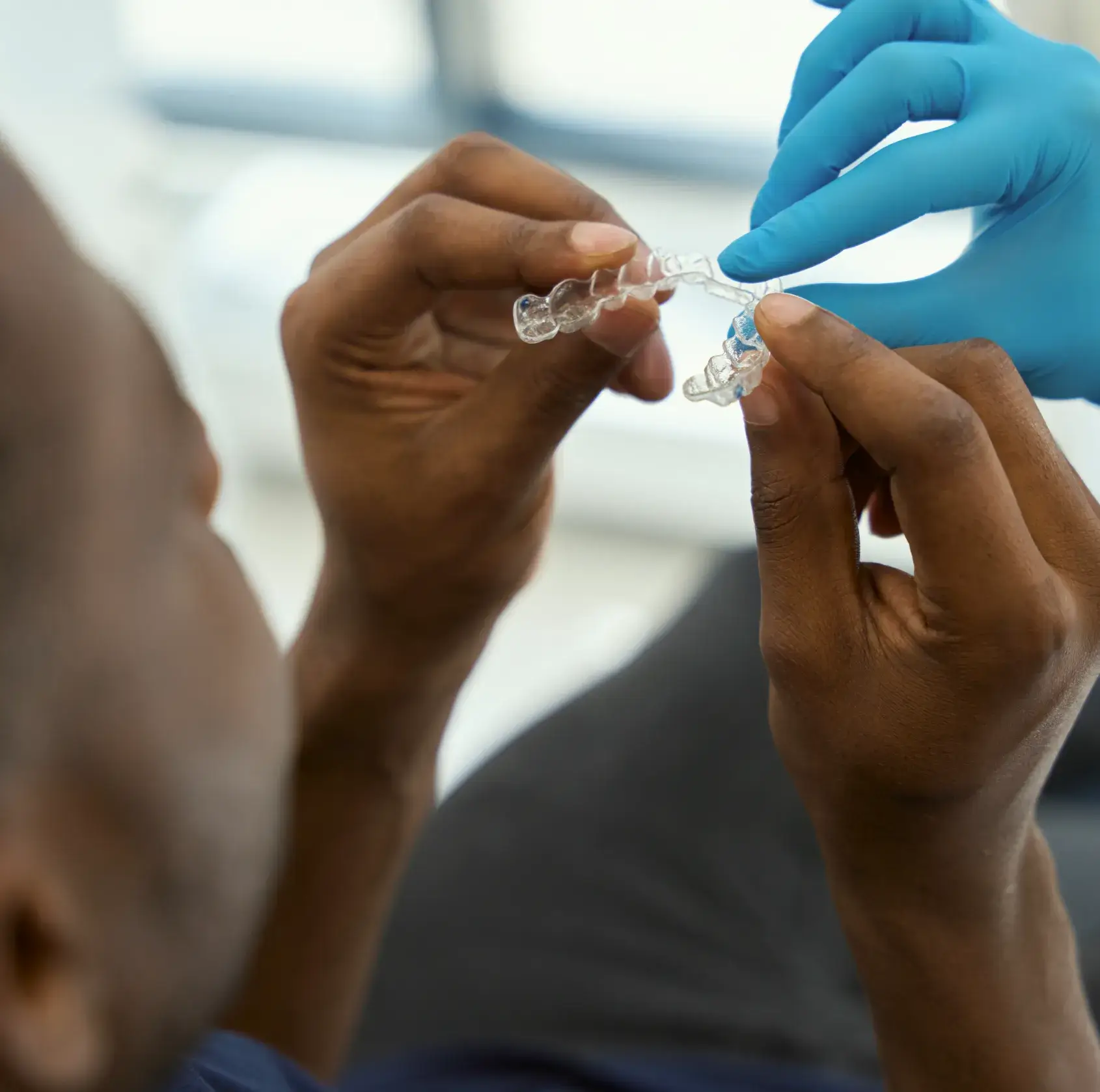 A dental professional wearing blue gloves showing a clear aligner to a patient holding it with both hands.