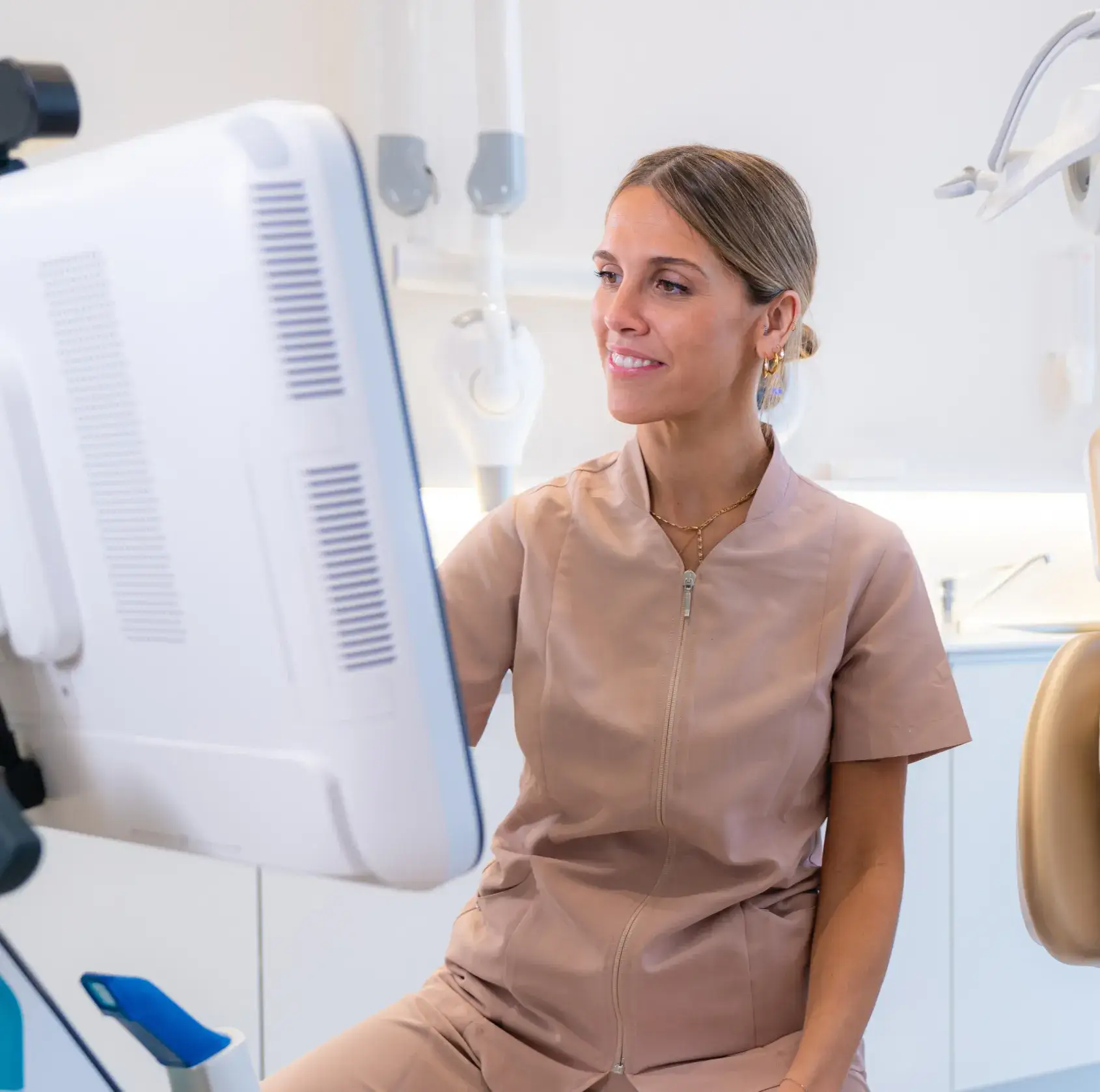 Female dental professional in beige scrubs looking at a computer screen in a dental clinic.