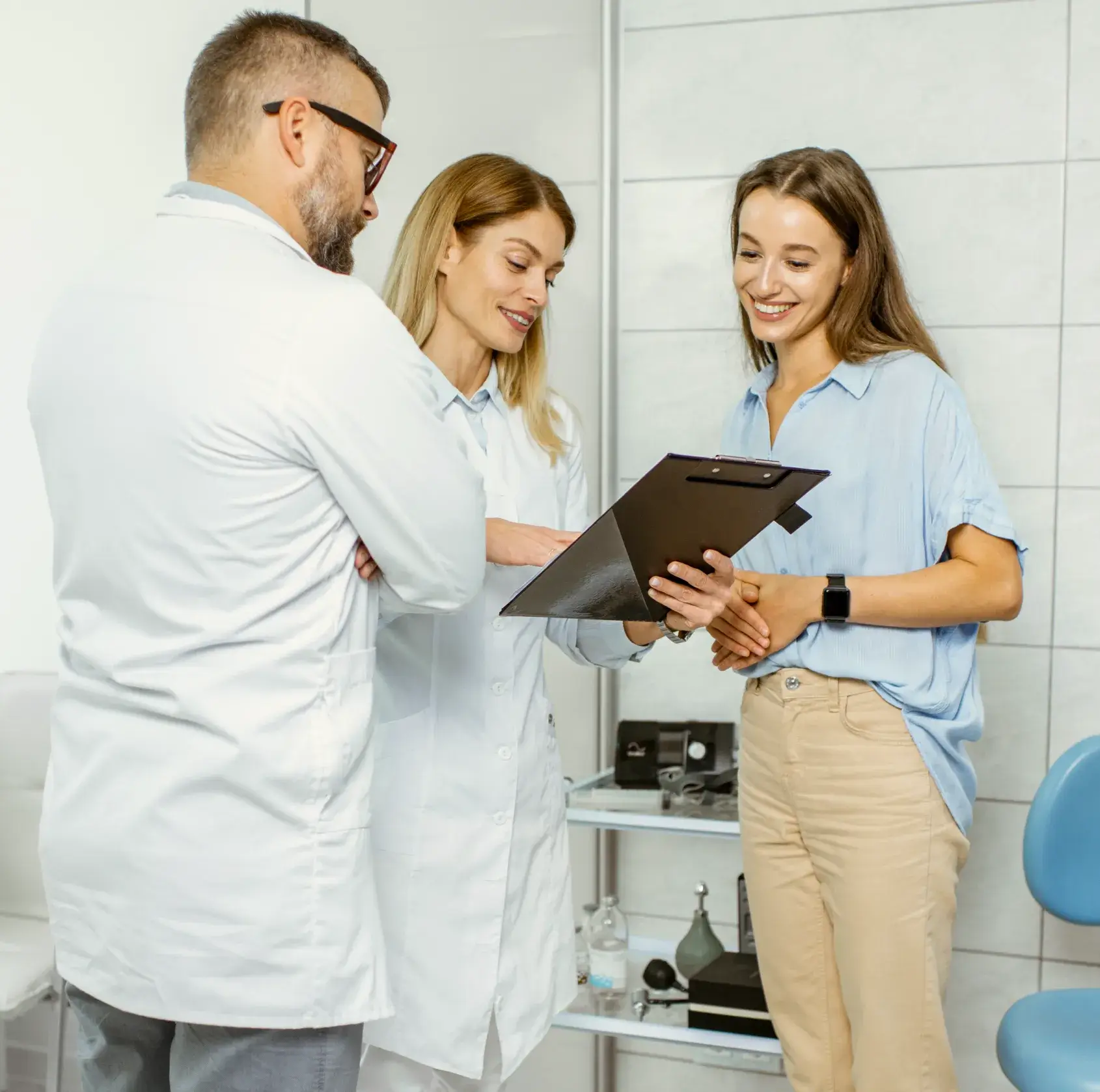 Two medical professionals in white coats discussing a clipboard with a smiling female patient in a light blue shirt and beige pants.